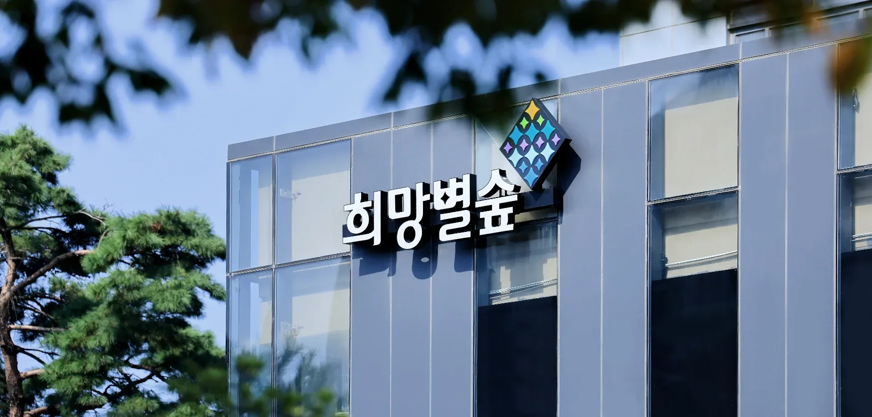 나무 사이로 보이는 희망별숲 건물 외벽의 흰 글자 간판과 여러 색의 별 모양 심벌.